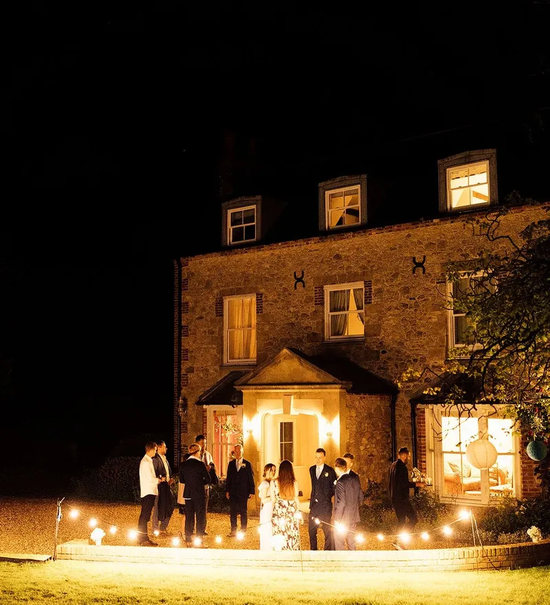 Guests gathered outside a lit stone house at night with string lights and paper lanterns