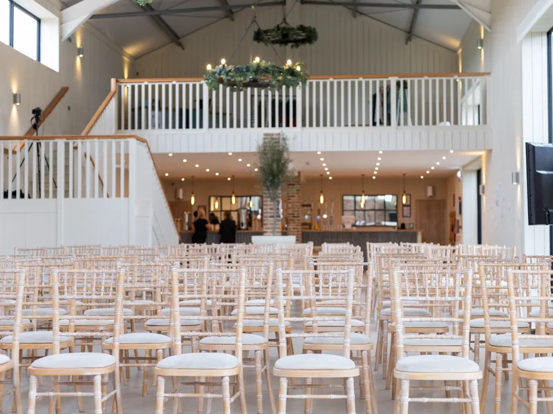 Rows of pale wooden chairs in a bright barn-style wedding venue with mezzanine balcony and bar