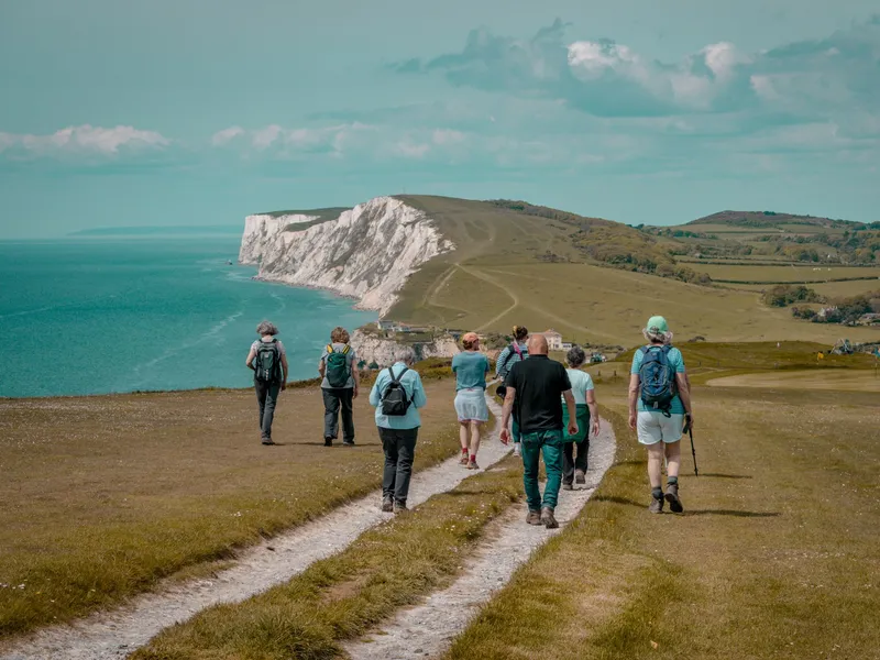 Group of hikers walking along a coastal path towards white chalk cliffs above a turquoise sea