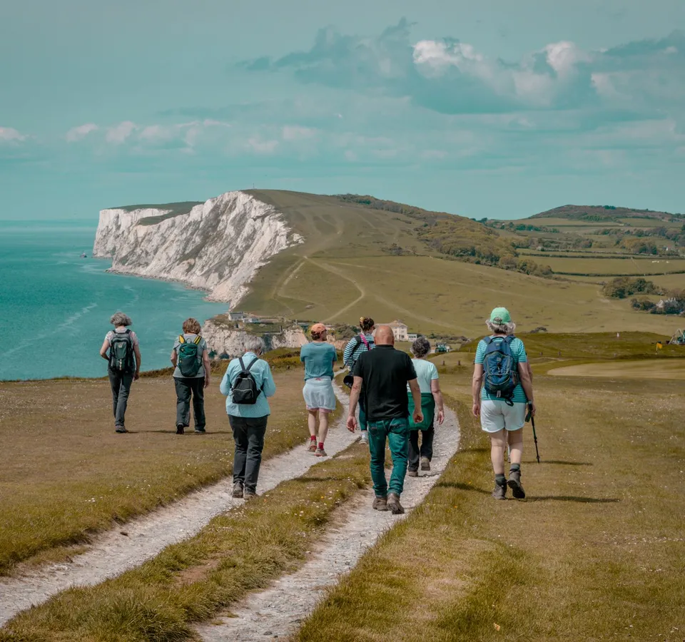 Group of hikers walking along a coastal path towards white chalk cliffs above a turquoise sea