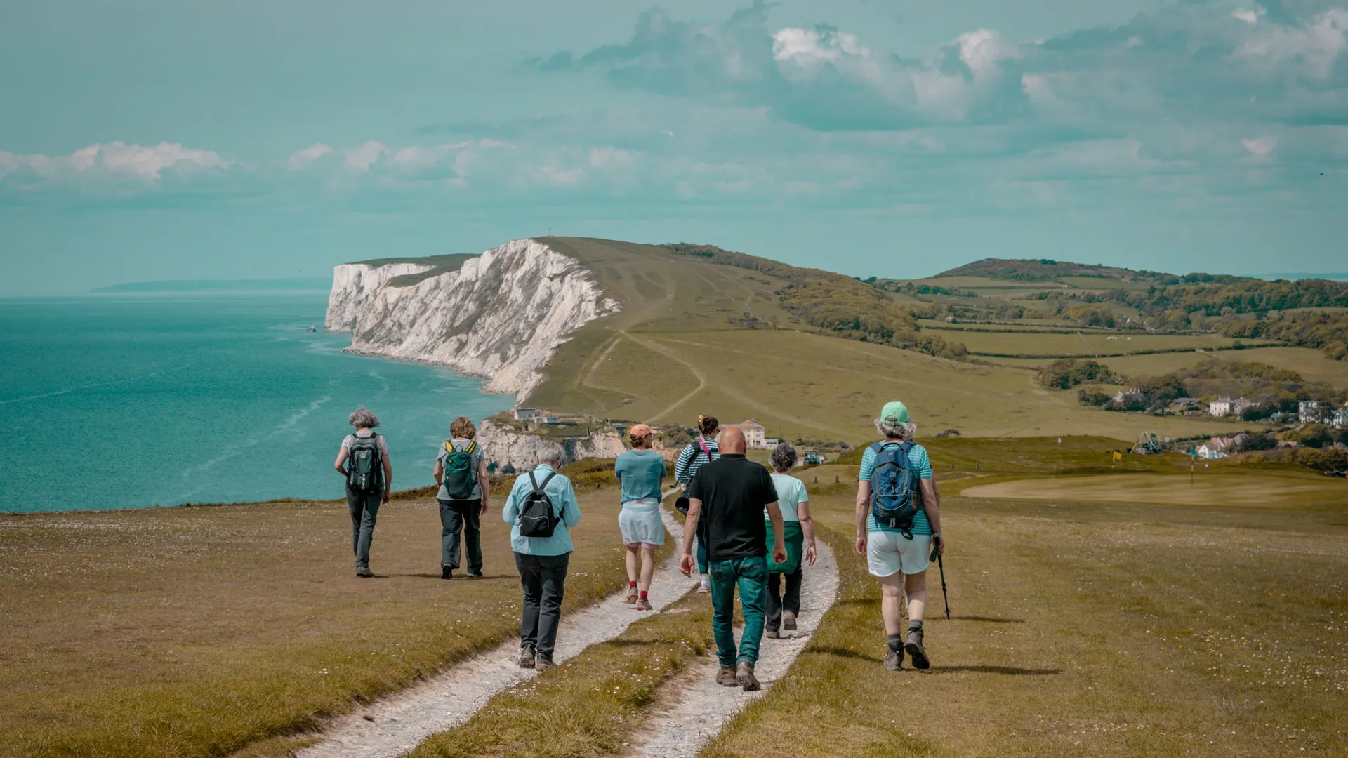 Group of hikers walking along a coastal path towards white chalk cliffs above a turquoise sea