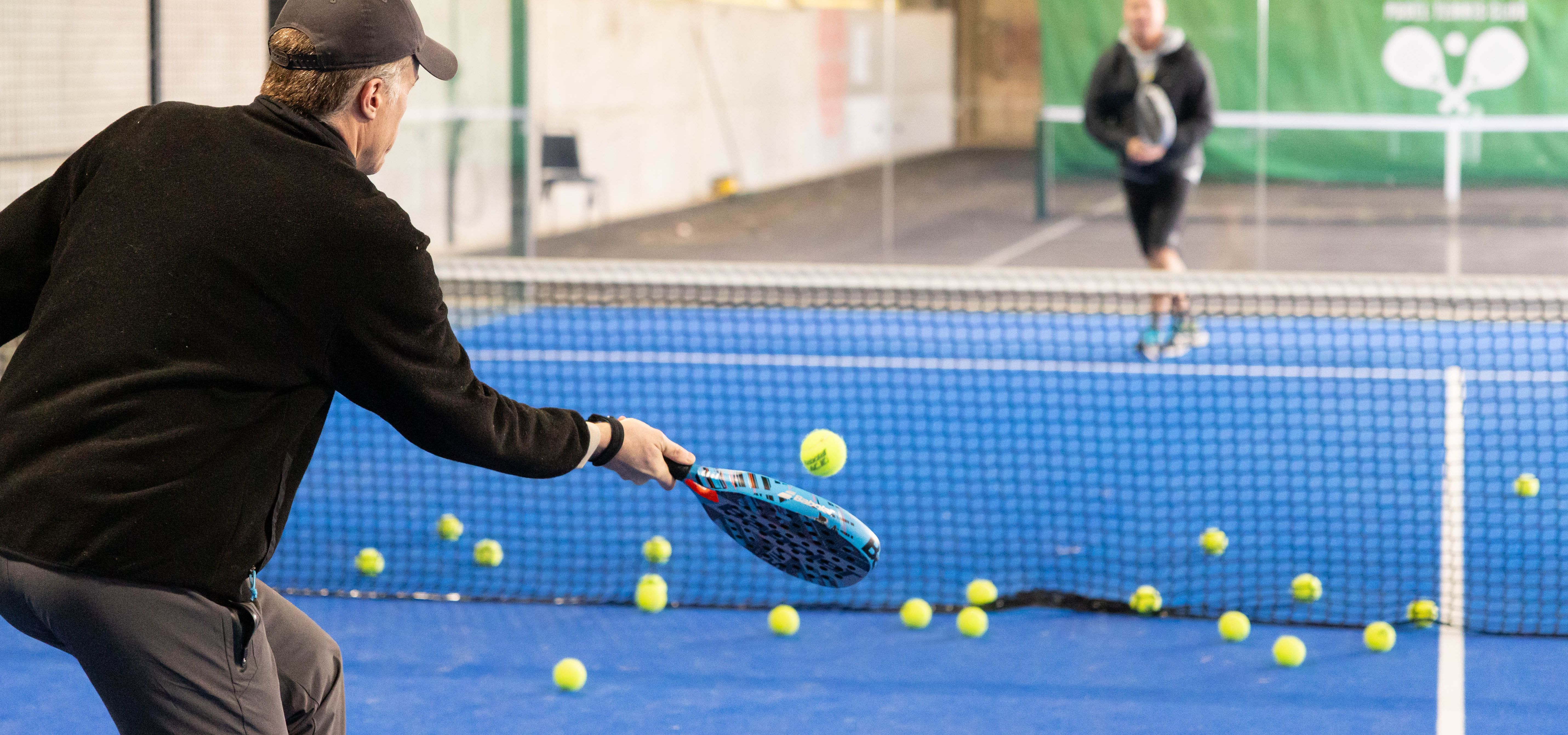 Man practising tennis volley at an indoor court with scattered balls and another player in the background