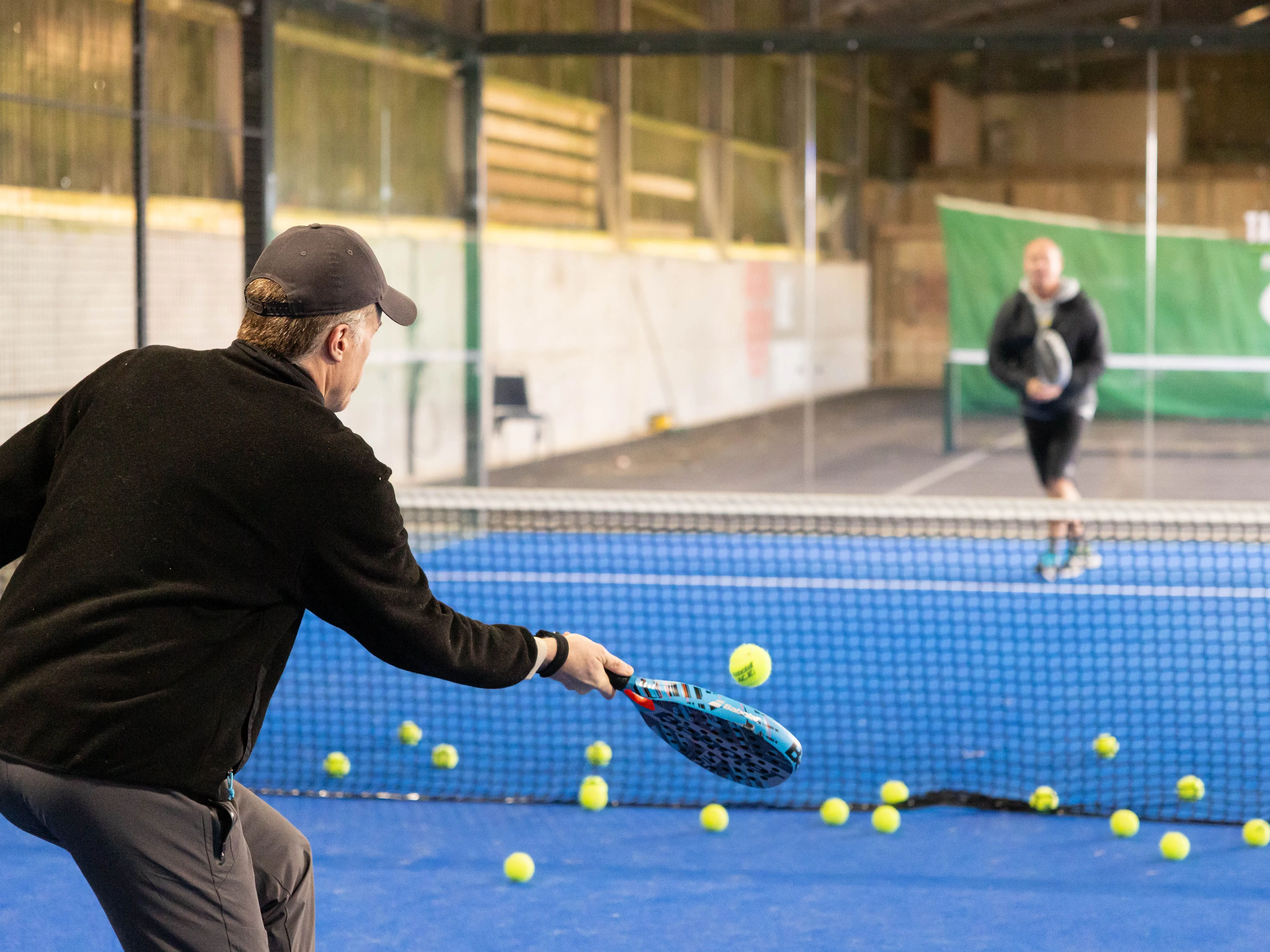 Man practising tennis volley at an indoor court with scattered balls and another player in the background