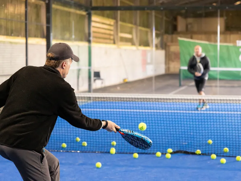 Man practising tennis volley at an indoor court with scattered balls and another player in the background