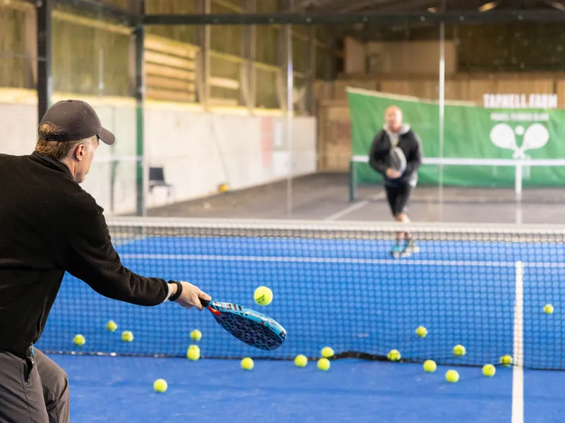 Man practising tennis volley at an indoor court with scattered balls and another player in the background