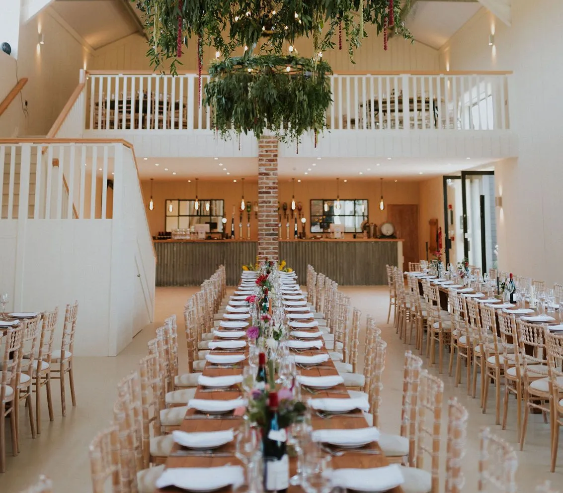 Long banquet table set for a celebration in a white barn hall with hanging greenery chandelier
