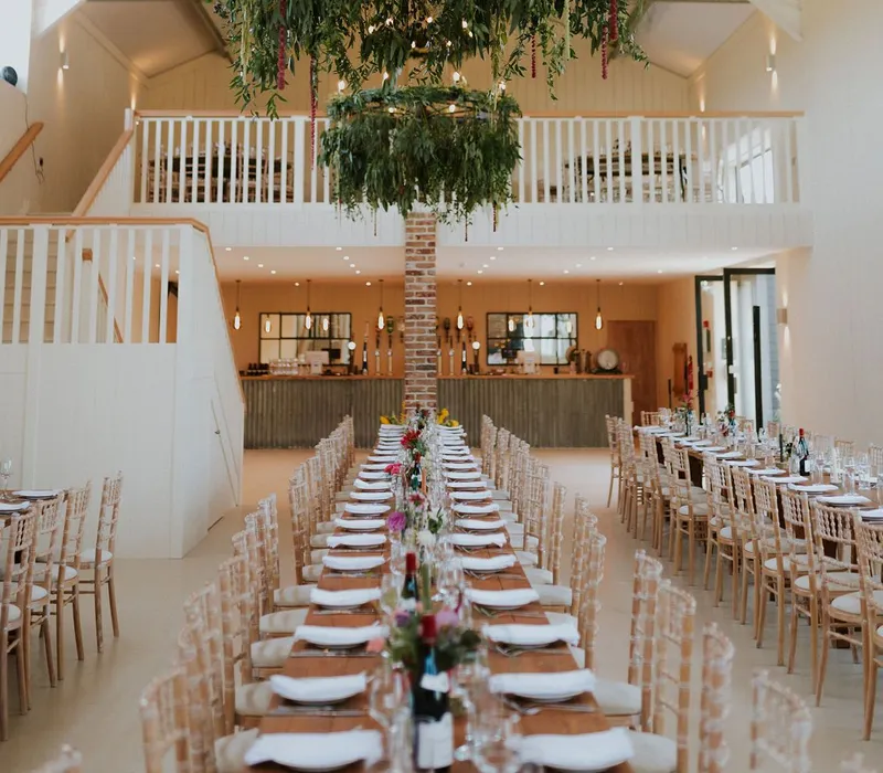 Long banquet table set for a celebration in a white barn hall with hanging greenery chandelier