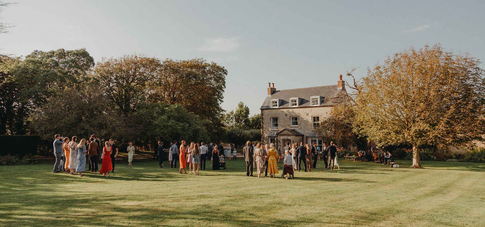 Wedding guests gathered on a lawn outside a large country house at sunset