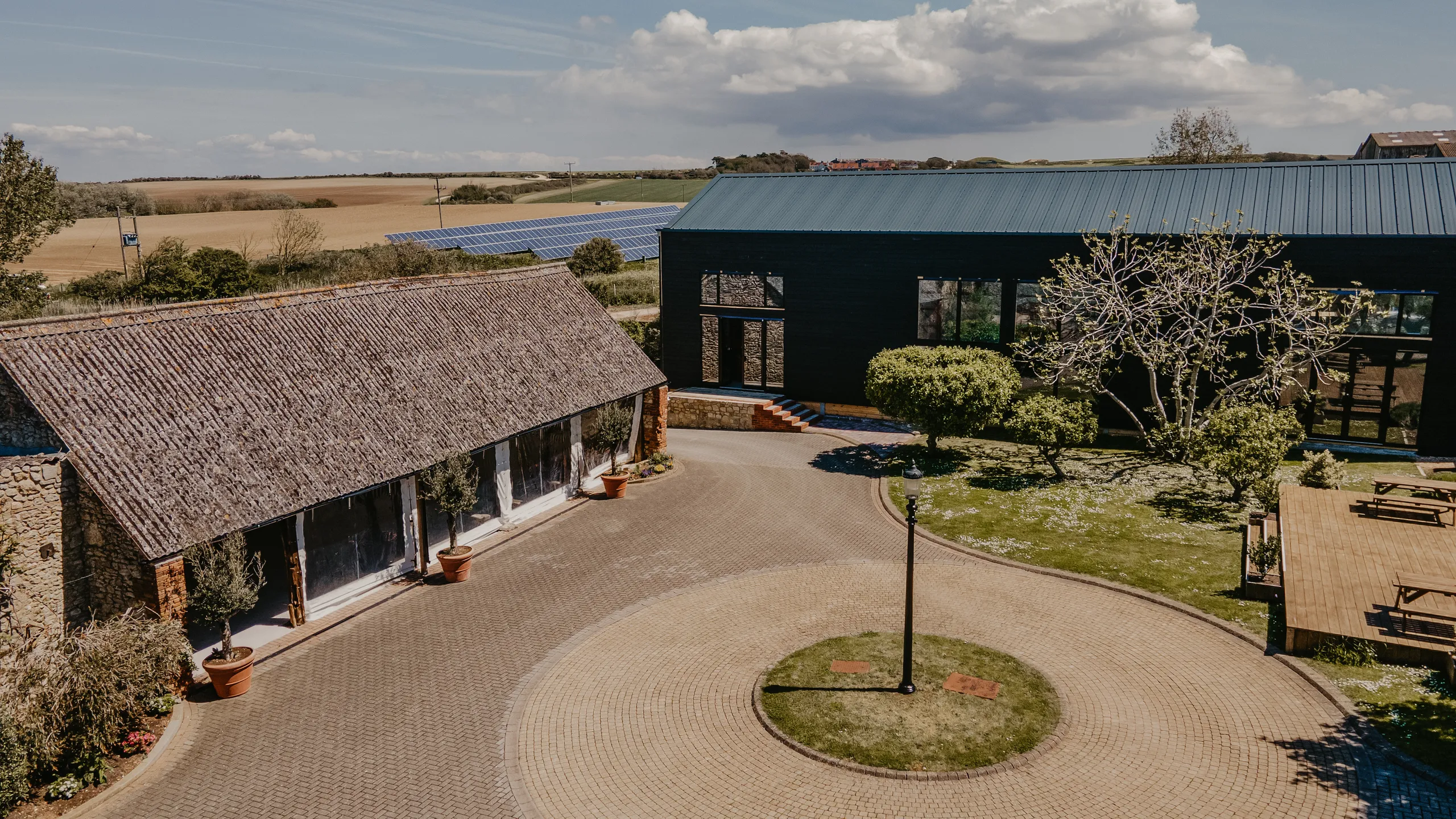 Aerial view of a rural courtyard with two barn-style buildings, a circular driveway, and solar panels in nearby fields