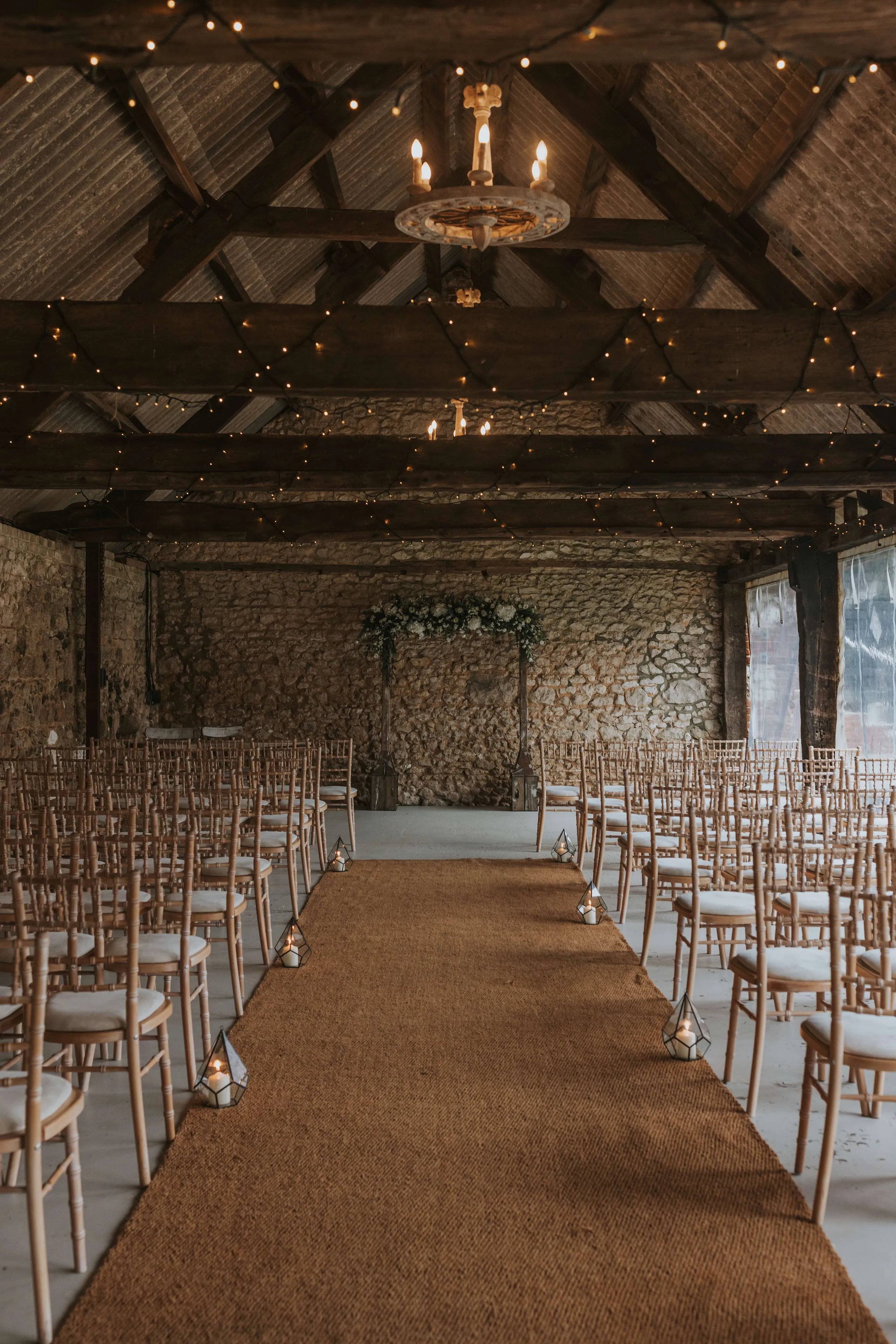 Rustic barn wedding ceremony set-up with aisle runner, lanterns and wooden chairs beneath fairy lights