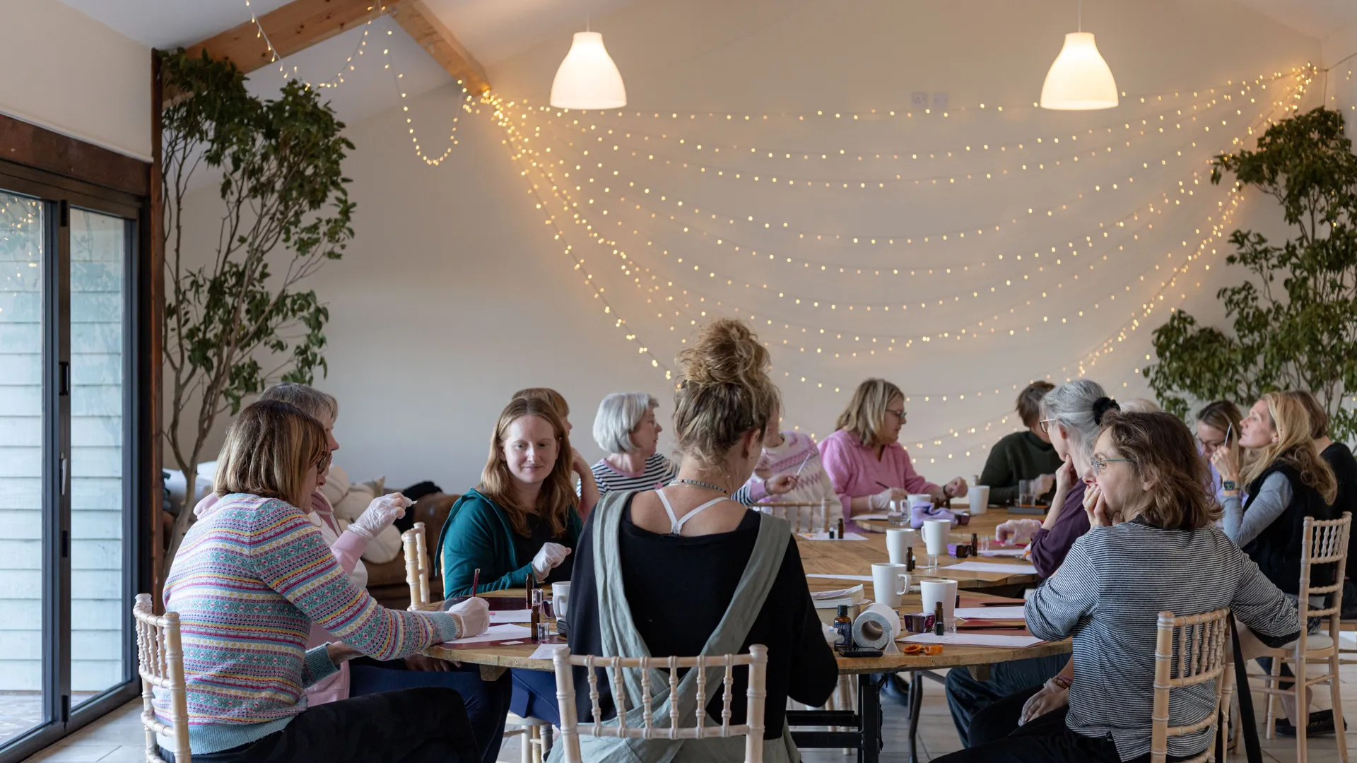 Group of women seated around tables in a bright room with fairy lights, talking and working on crafts