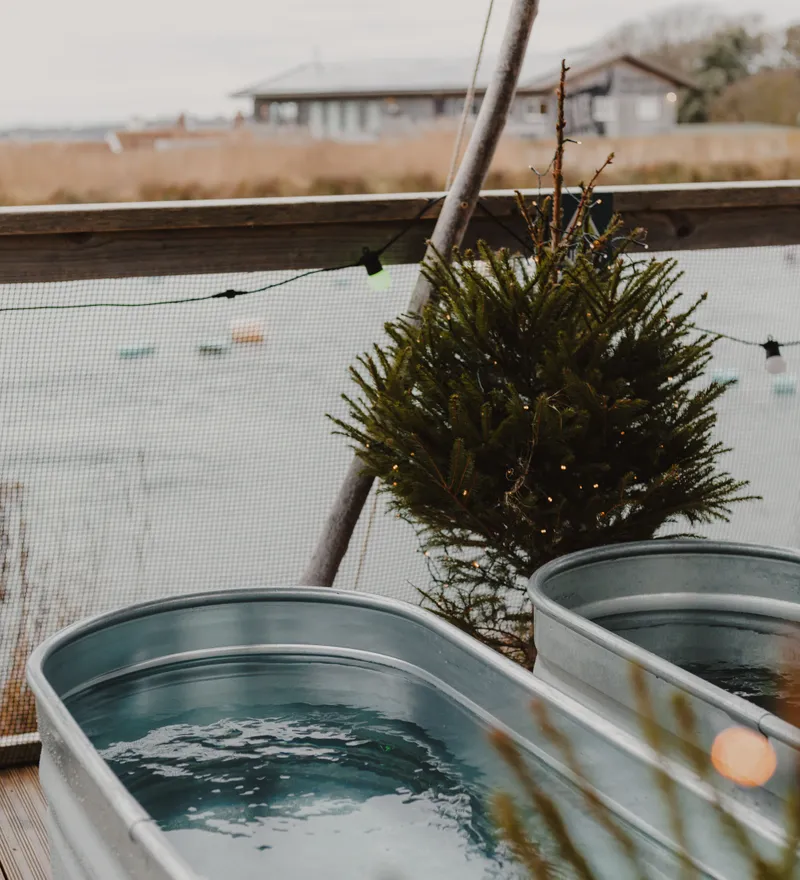 Two metal tubs filled with water on a wooden deck overlooking a lake, with a small fir tree and string lights nearby