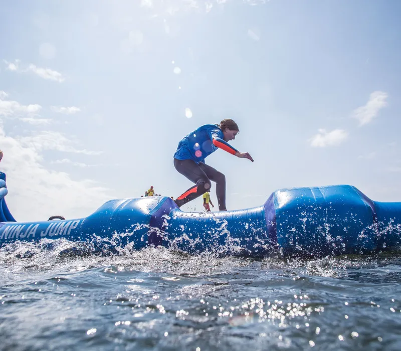 Participants run across a blue inflatable water obstacle course with splashing water under a bright sky