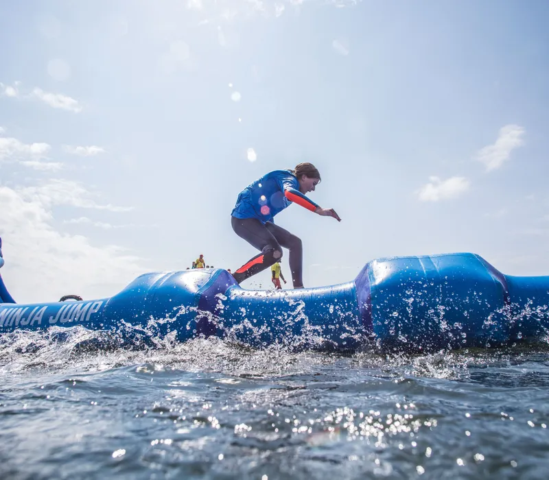 Participants run across a blue inflatable water obstacle course with splashing water under a bright sky