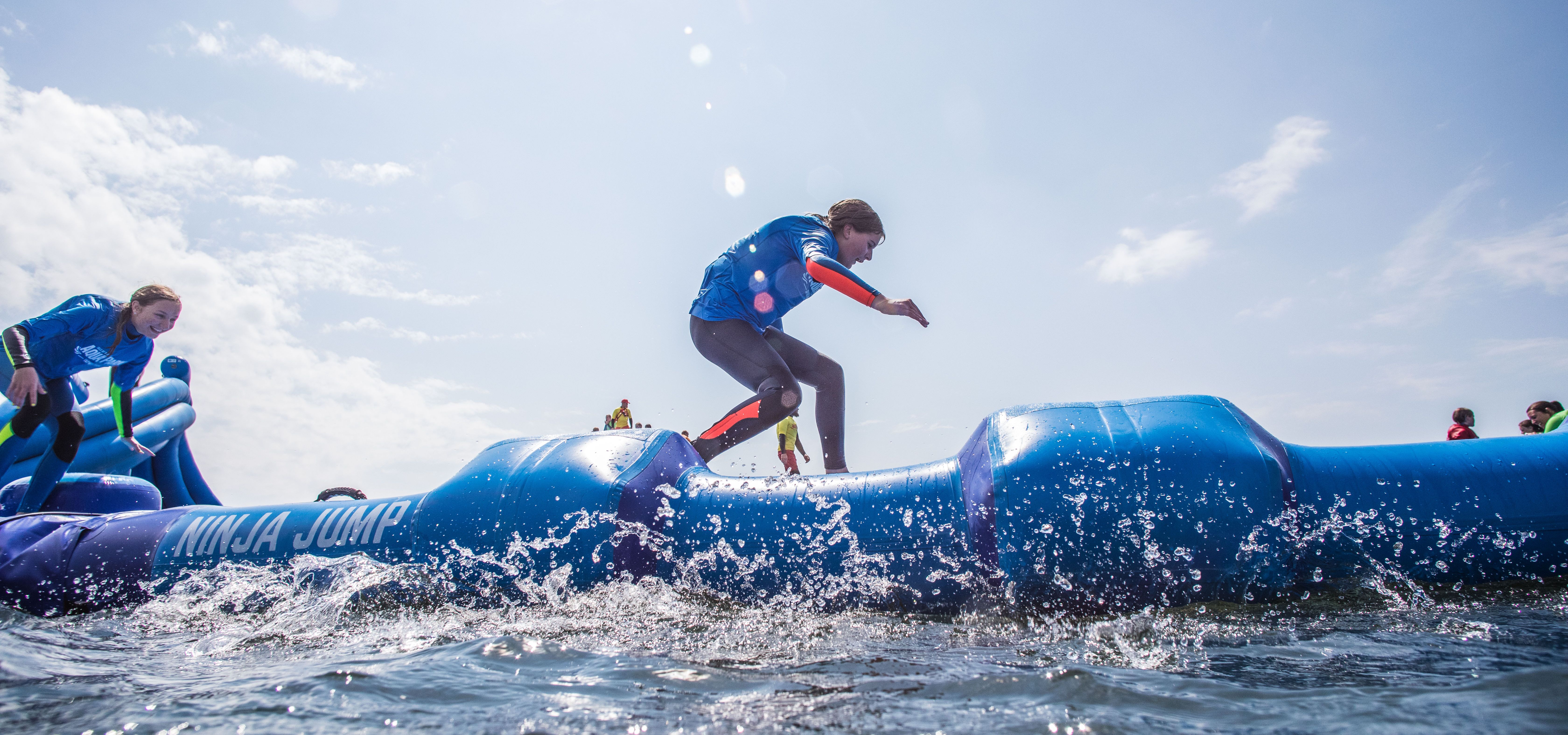 Participants run across a blue inflatable water obstacle course with splashing water under a bright sky