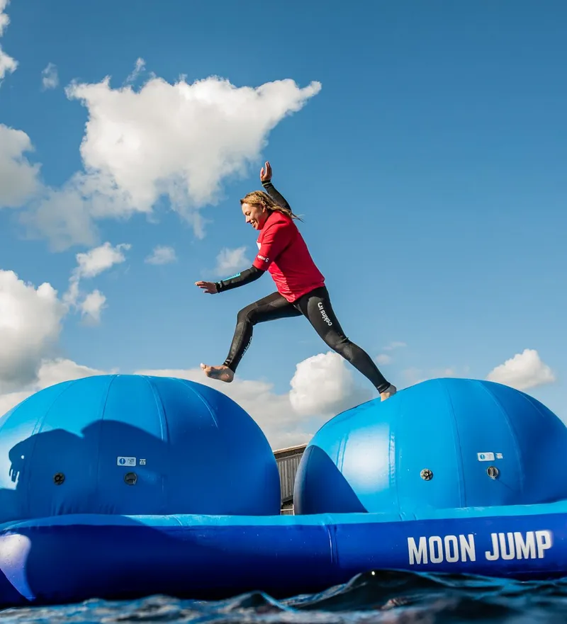 Person in a red top and wetsuit leaping between large blue inflatable platforms on the water under a cloudy blue sky