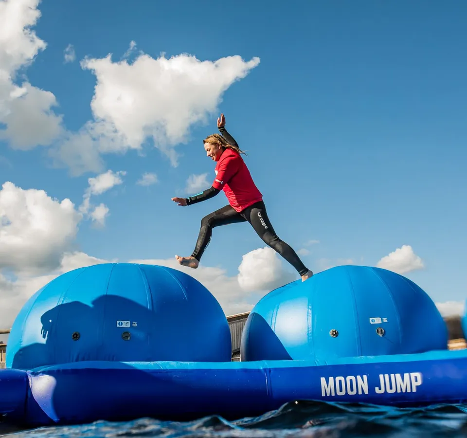 Person in a red top and wetsuit leaping between large blue inflatable platforms on the water under a cloudy blue sky