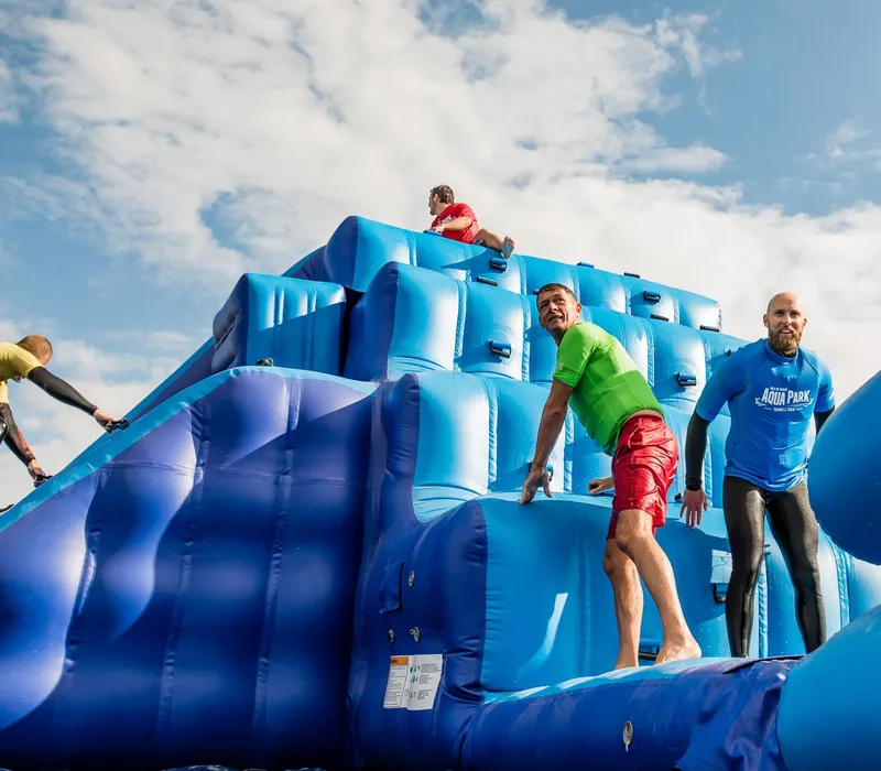 People climbing a large blue inflatable obstacle at an outdoor aqua park under a cloudy sky