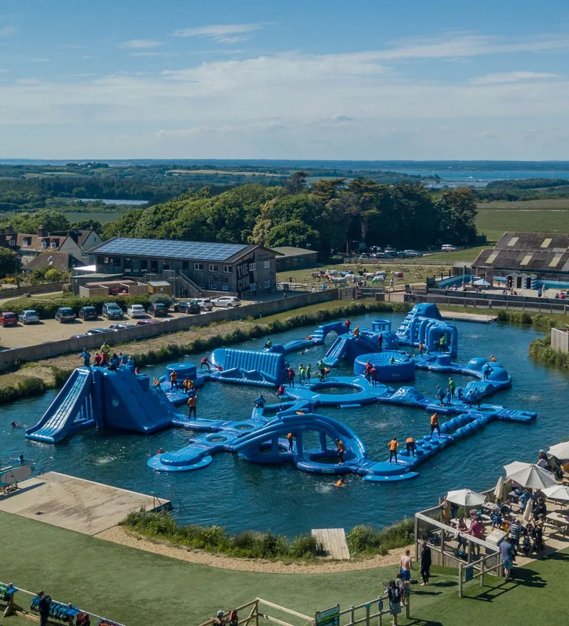 Aerial view of a large blue inflatable water obstacle course in a lake with people climbing and sliding, beside farm buildings and parked cars.