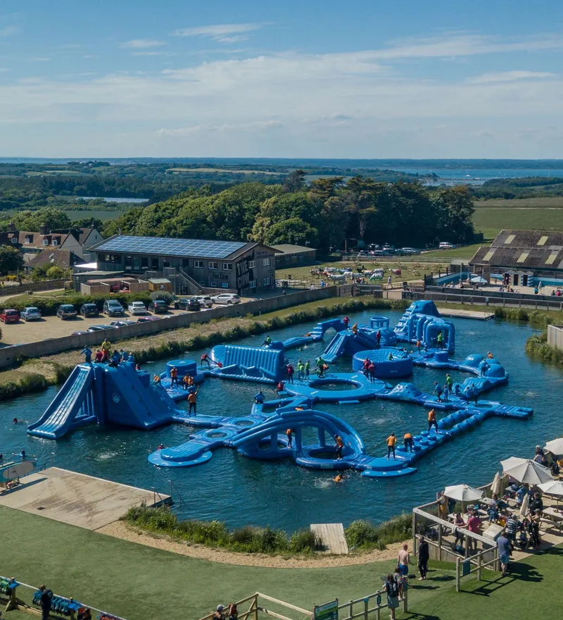 Aerial view of a large blue inflatable water obstacle course in a lake with people climbing and sliding, beside farm buildings and parked cars.