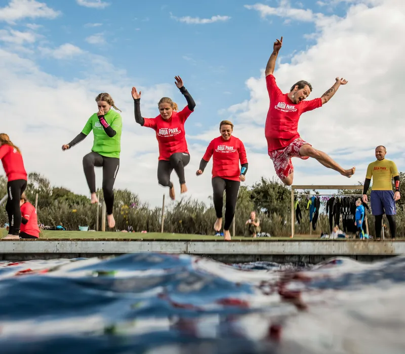 People in wetsuits jump from a dock into open water at an outdoor aqua park
