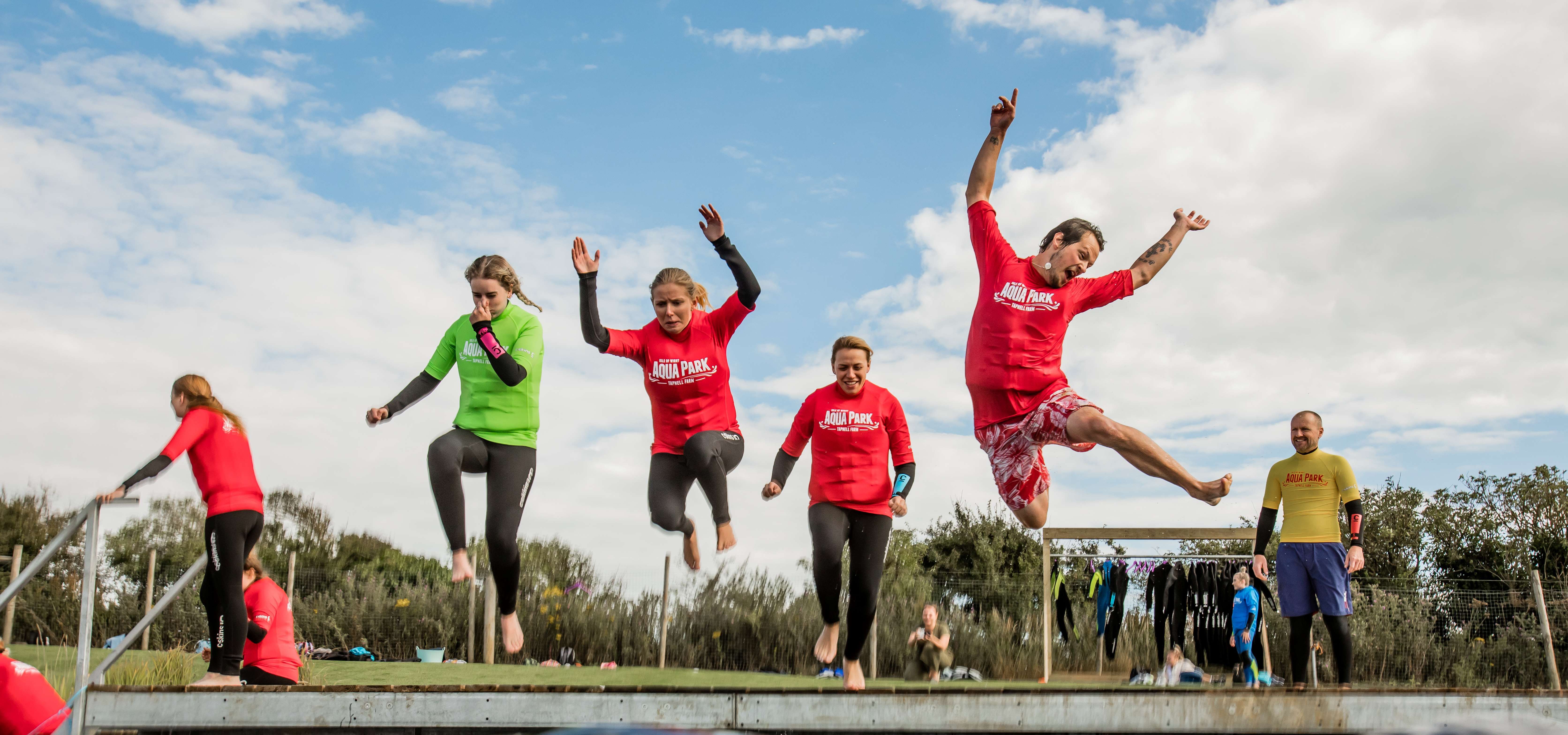 People in wetsuits jump from a dock into open water at an outdoor aqua park