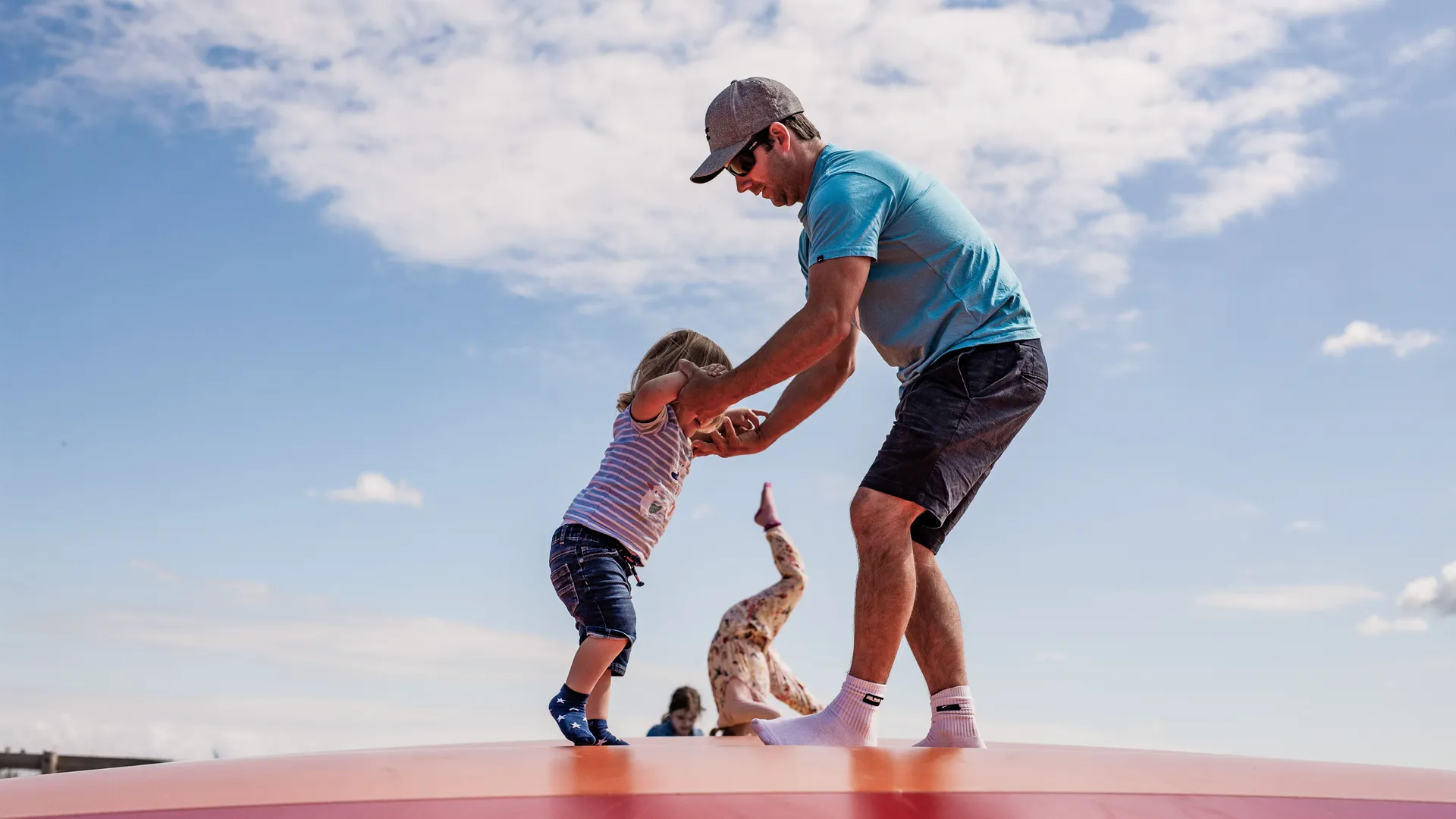 Adult helping a young child balance on an inflatable bouncy pillow outdoors under a blue sky