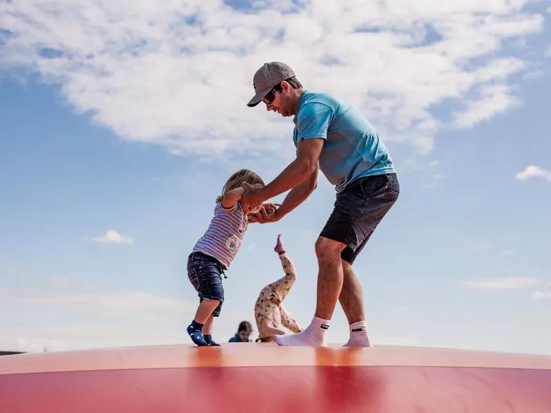 Adult helping a young child balance on an inflatable bouncy pillow outdoors under a blue sky