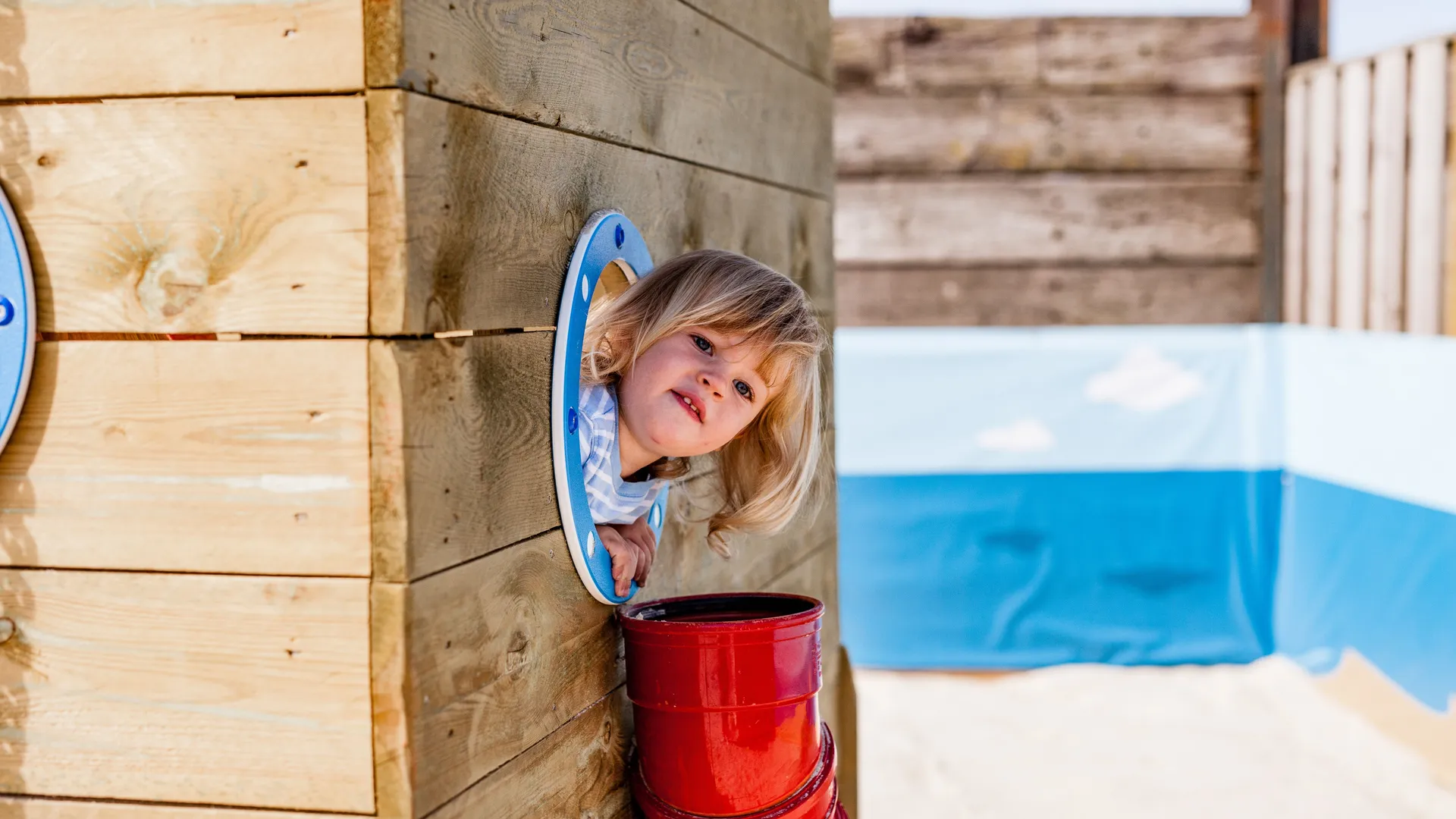 Young child peeking through a round window in a wooden play structure at the beach