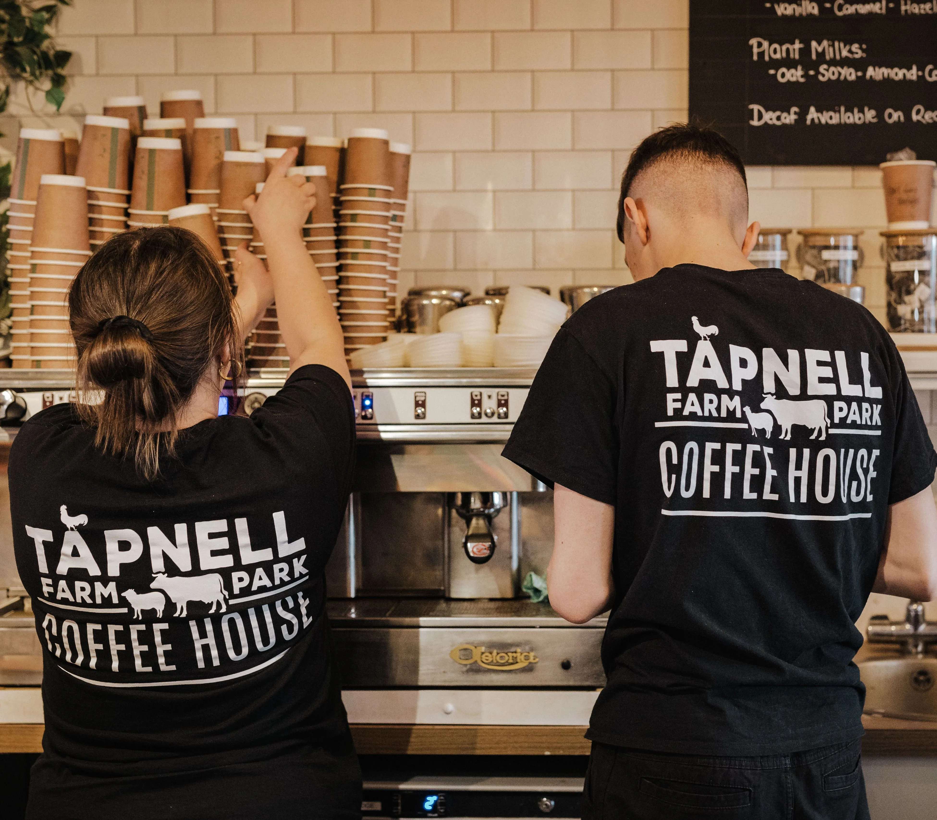 Two coffee shop staff in black Tapnell Farm Park Coffee House T-shirts working at an espresso machine behind a counter.