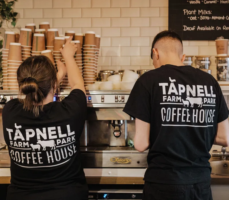 Two coffee shop staff in black Tapnell Farm Park Coffee House T-shirts working at an espresso machine behind a counter.
