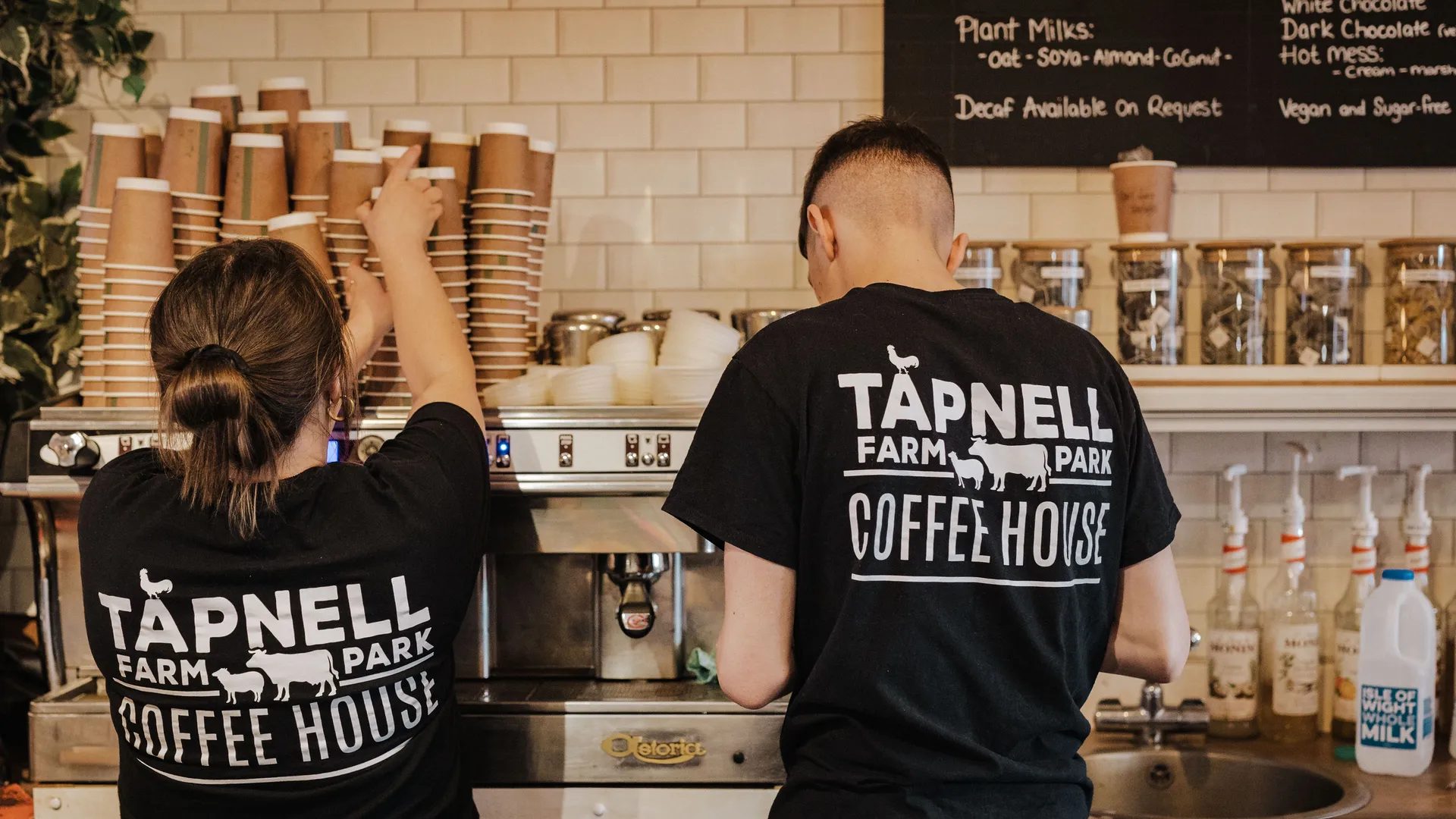 Two coffee shop staff in black Tapnell Farm Park Coffee House T-shirts working at an espresso machine behind a counter.