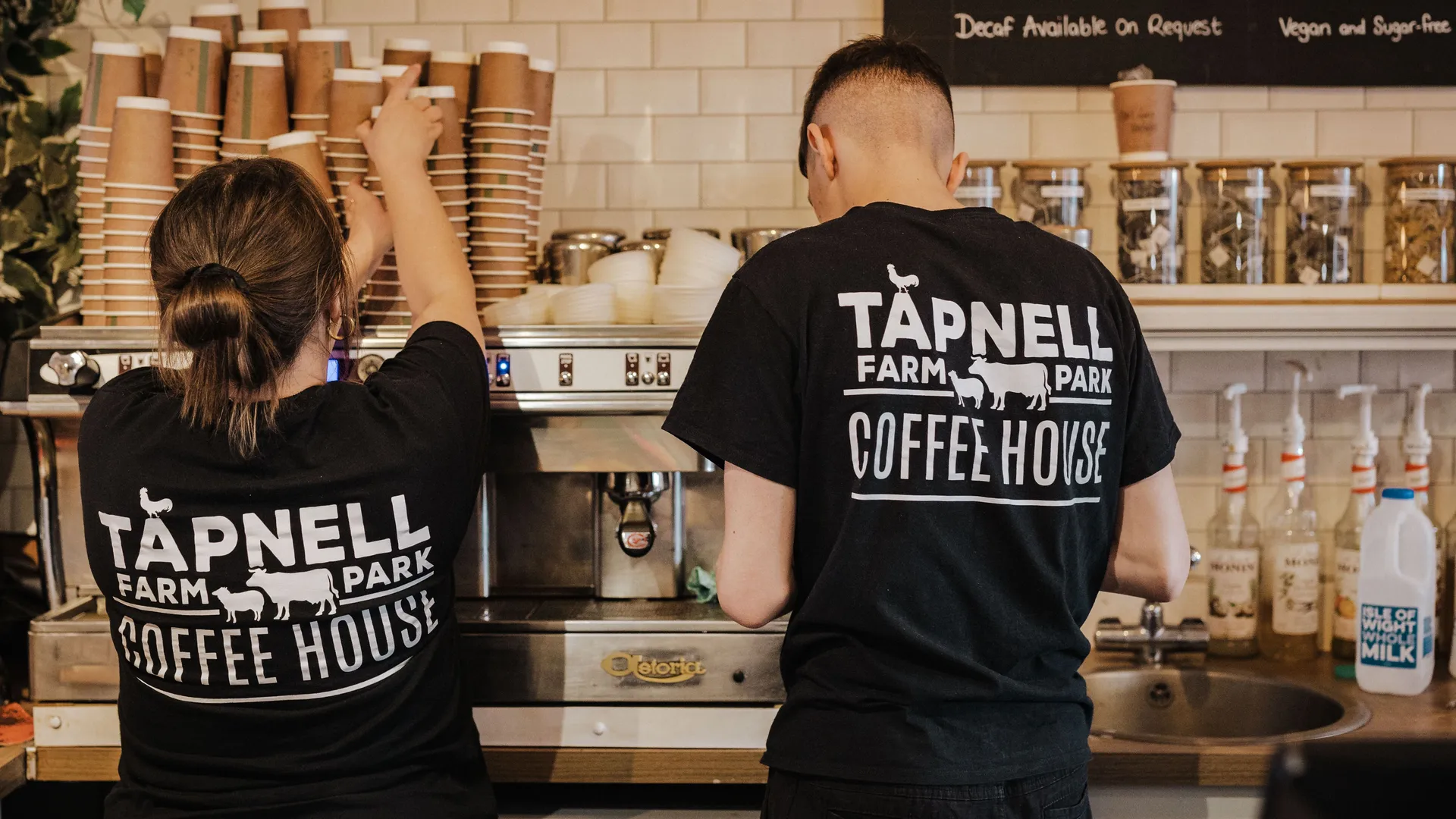 Two coffee shop staff in black Tapnell Farm Park Coffee House T-shirts working at an espresso machine behind a counter.