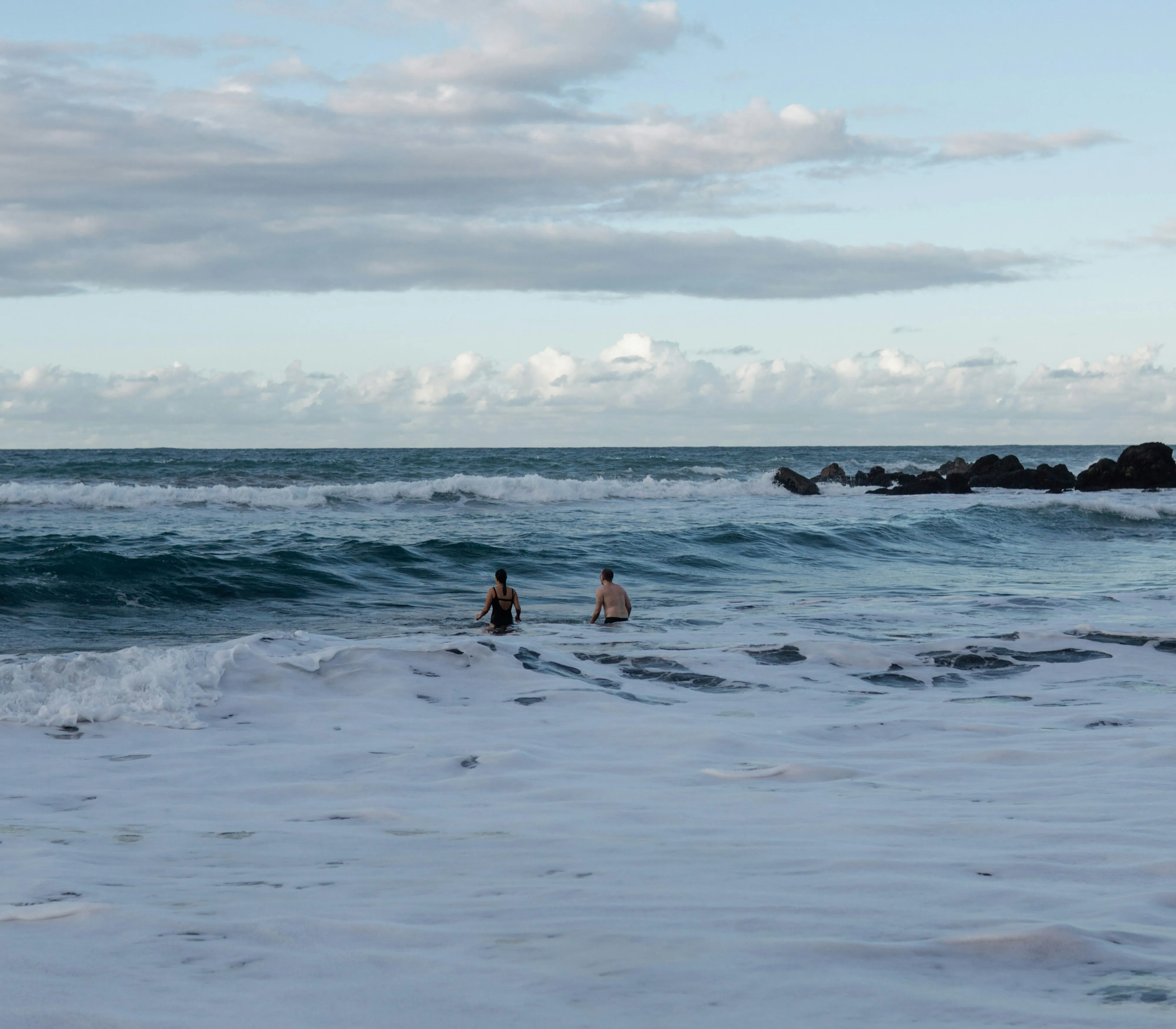 Two people wading in the sea as waves roll in under a cloudy blue sky