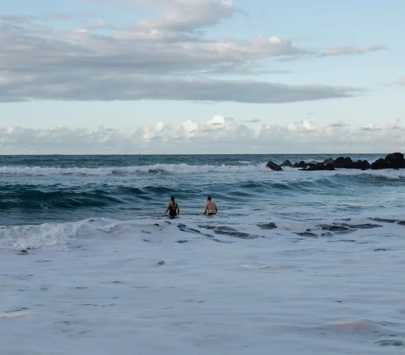 Two people wading in the sea as waves roll in under a cloudy blue sky