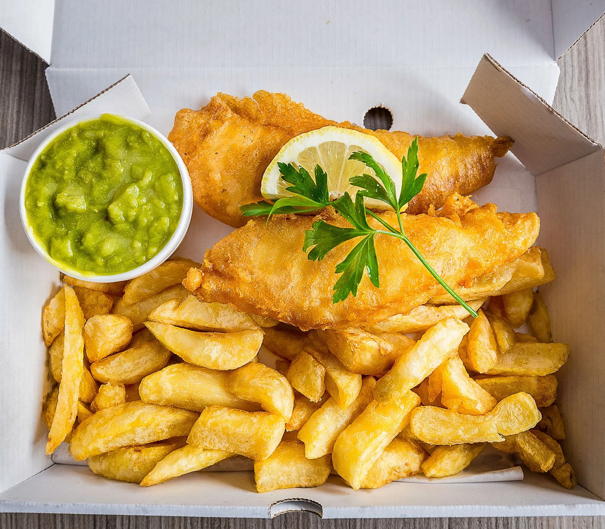Fish and chips in a takeaway box with mushy peas, lemon and parsley