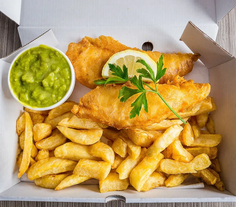 Fish and chips in a takeaway box with mushy peas, lemon and parsley