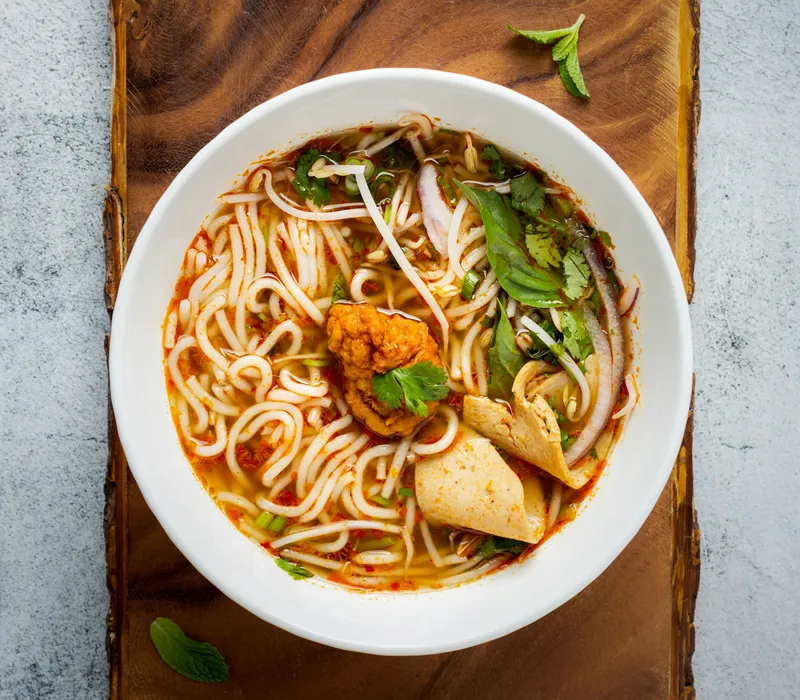 Bowl of spicy noodle soup with herbs and sliced onion on a wooden board