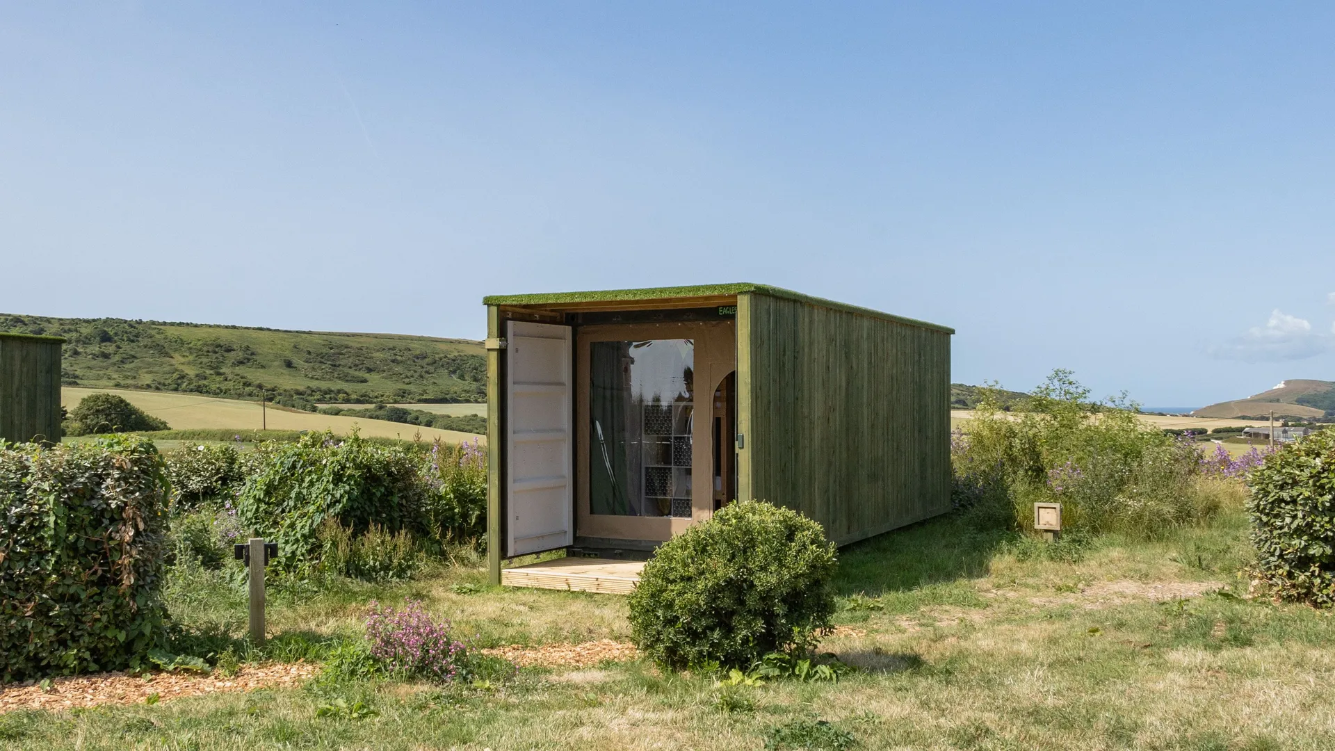 Green wooden container cabin with open door in a grassy field with rolling hills under a clear blue sky