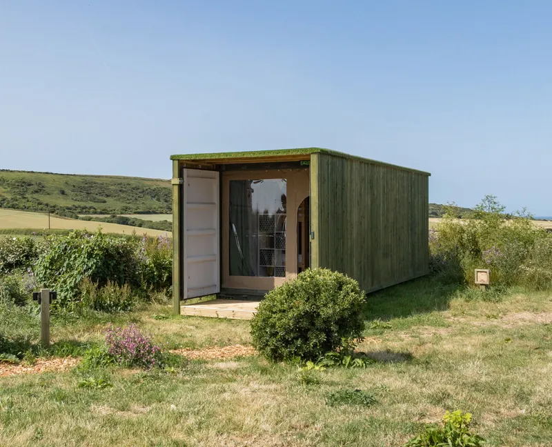 Green wooden container cabin with open door in a grassy field with rolling hills under a clear blue sky