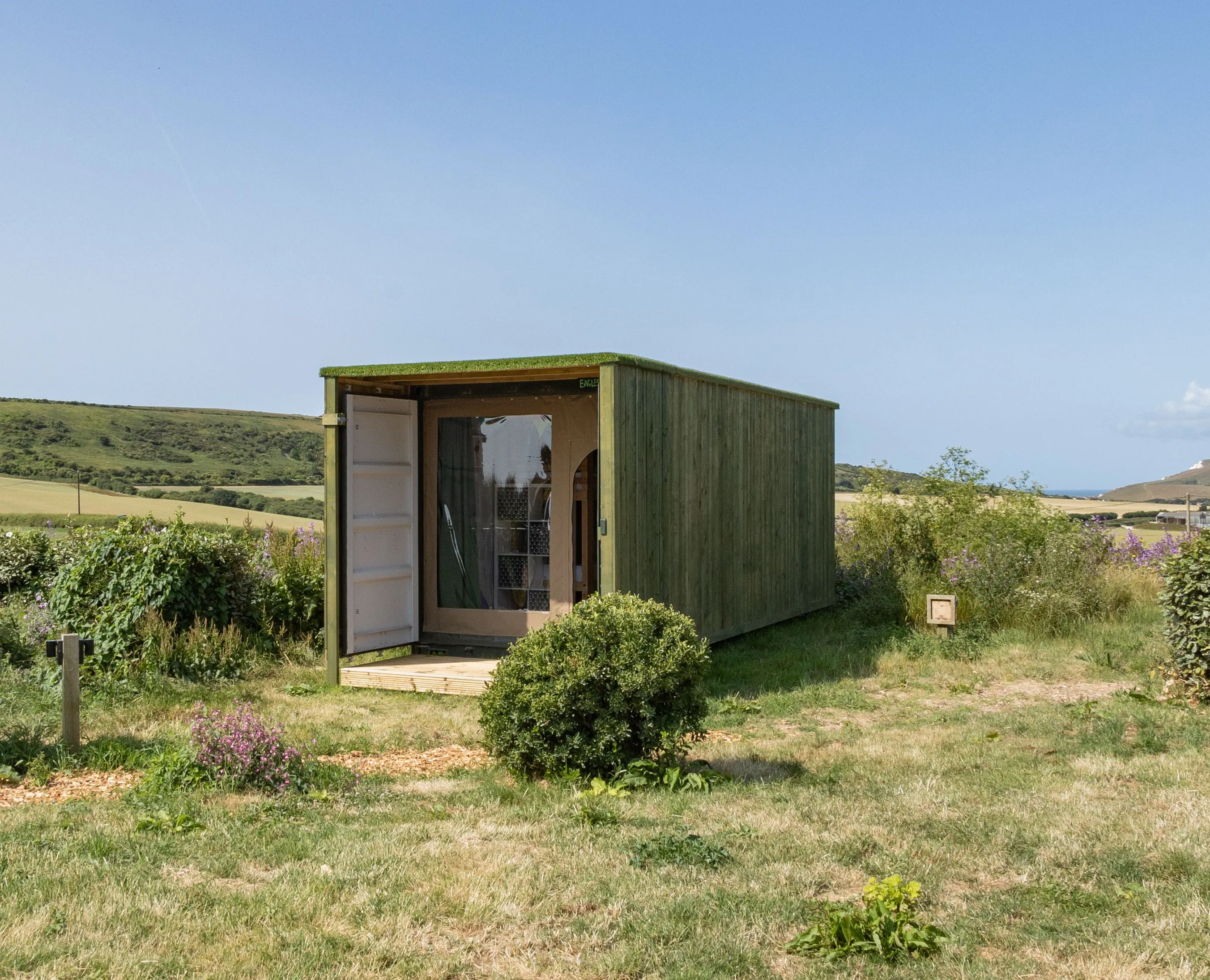 Green wooden container cabin with open door in a grassy field with rolling hills under a clear blue sky