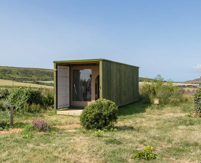Green wooden container cabin with open door in a grassy field with rolling hills under a clear blue sky