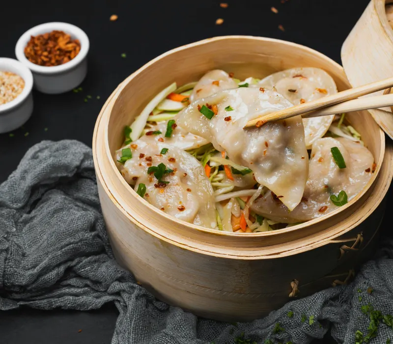 Chopsticks lifting a steamed dumpling from a bamboo basket with shredded vegetables and chilli flakes