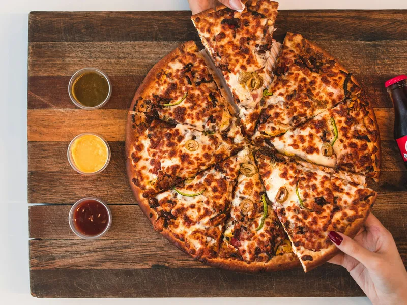 Two hands taking slices of cheesy pizza on a wooden board with three dipping sauces and a bottle of Coca-Cola
