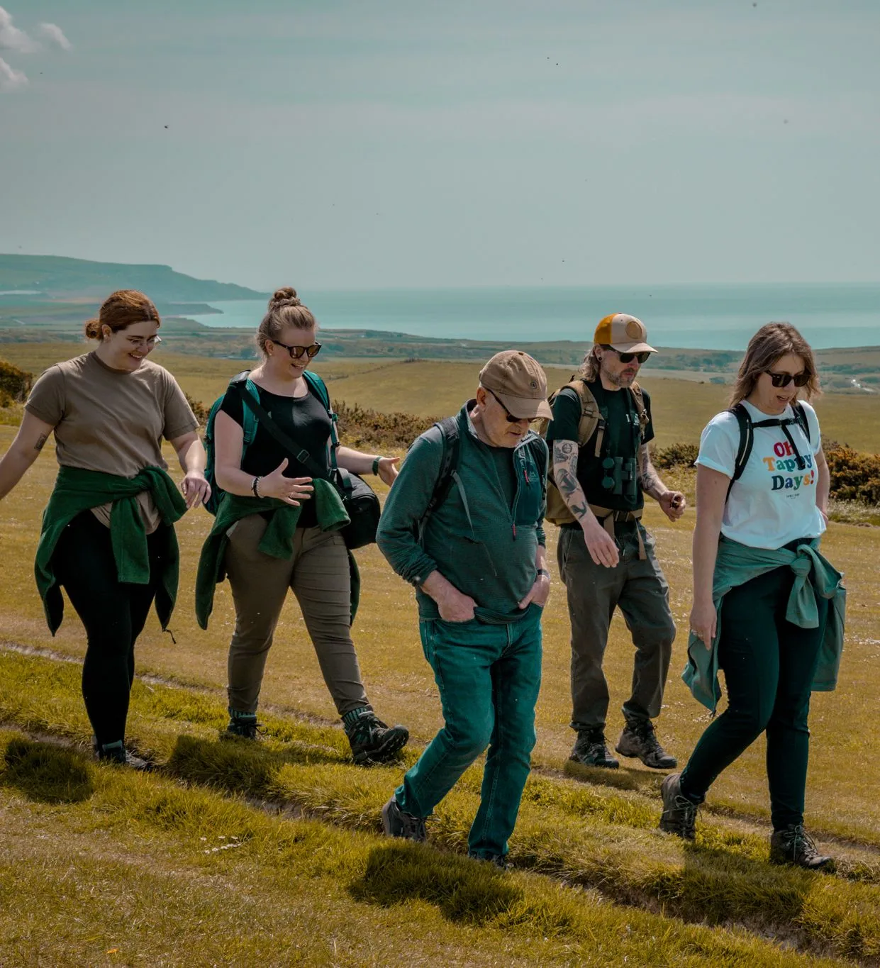 Group of five hikers walking along a grassy coastal hillside with the sea in the distance