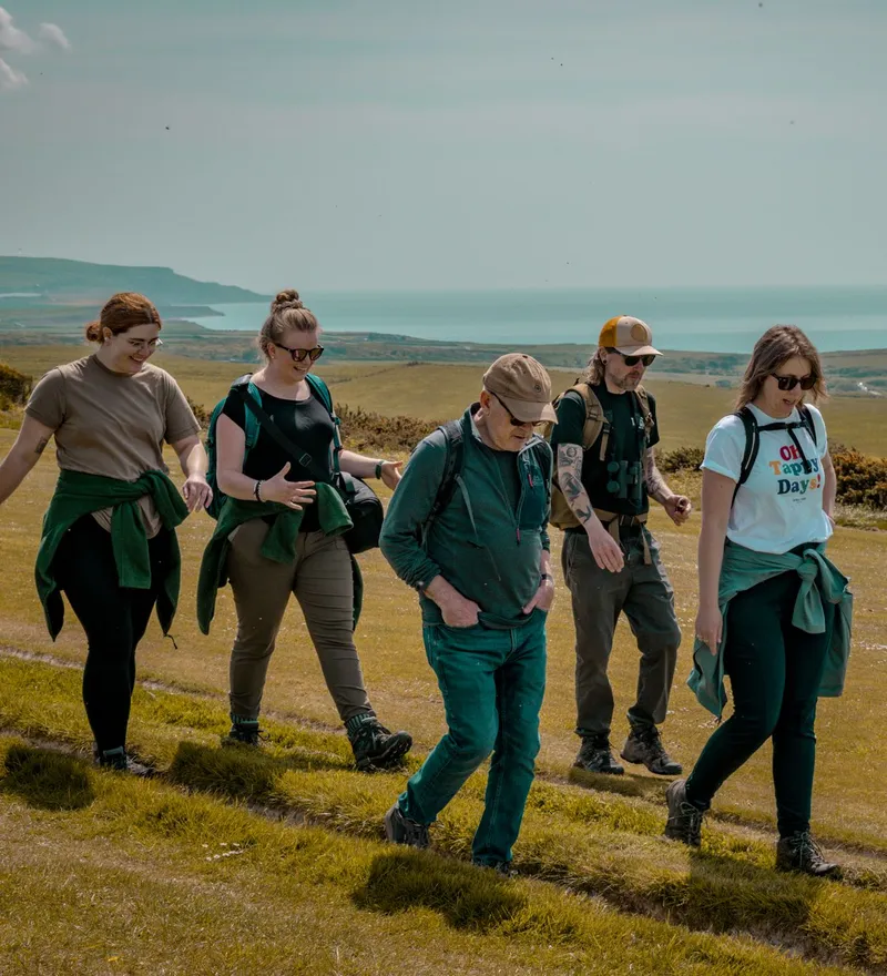 Group of five hikers walking along a grassy coastal hillside with the sea in the distance