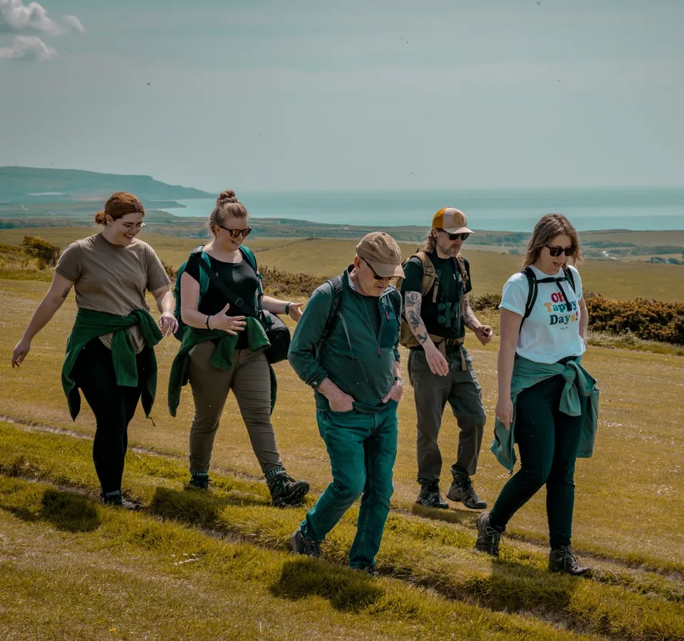 Group of five hikers walking along a grassy coastal hillside with the sea in the distance