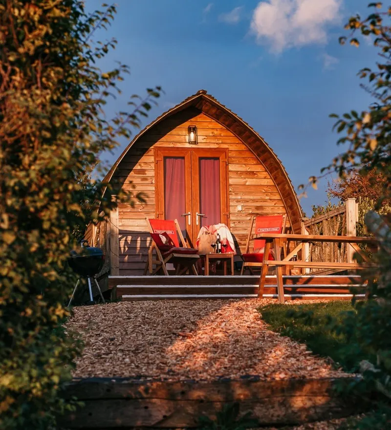 Wooden eco pod cabin with outdoor chairs and a BBQ, surrounded by greenery