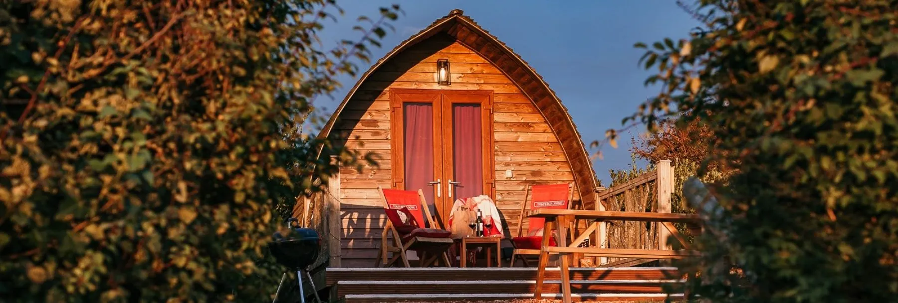 Wooden eco pod cabin with outdoor chairs and a BBQ, surrounded by greenery