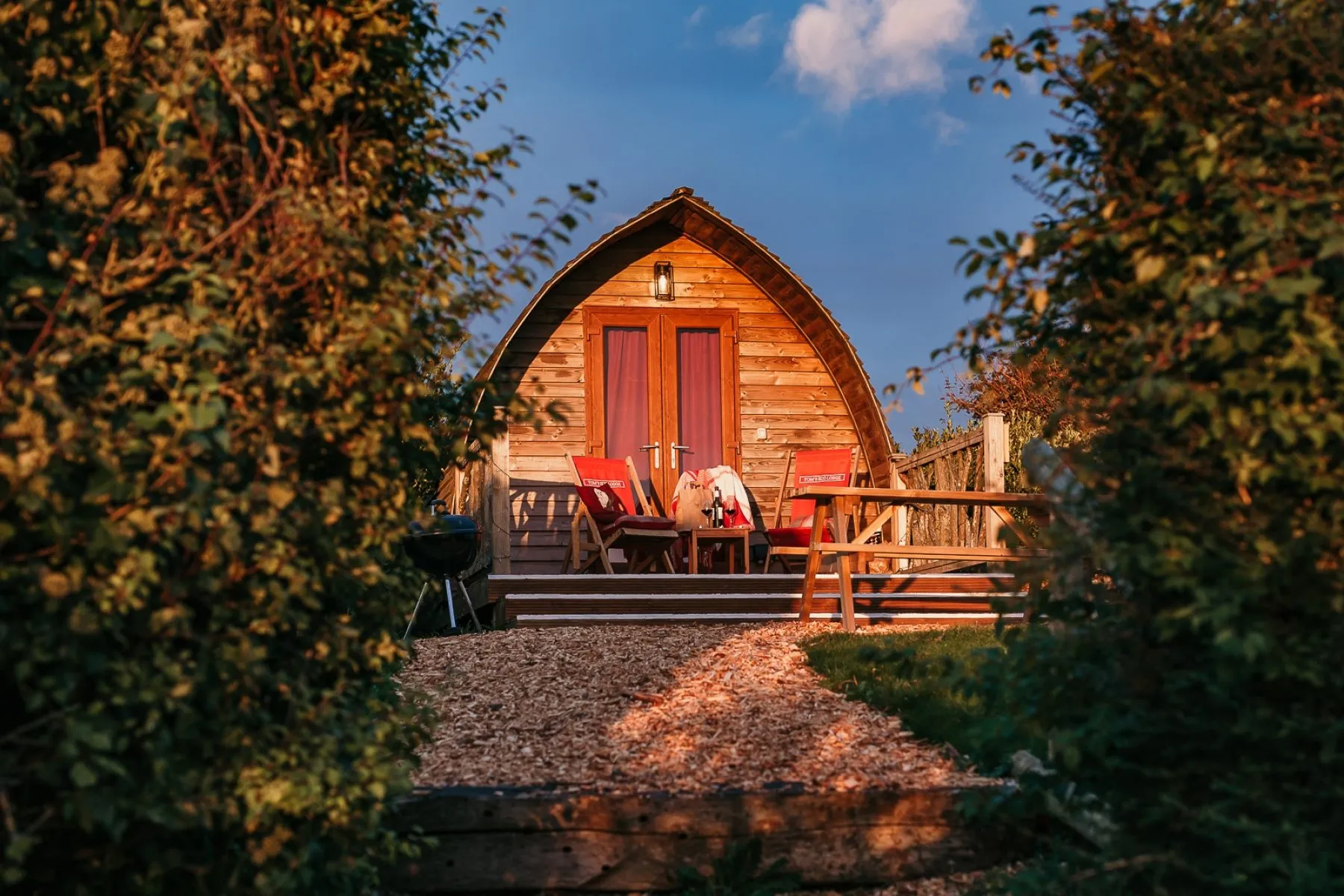Wooden eco pod cabin with outdoor chairs and a BBQ, surrounded by greenery